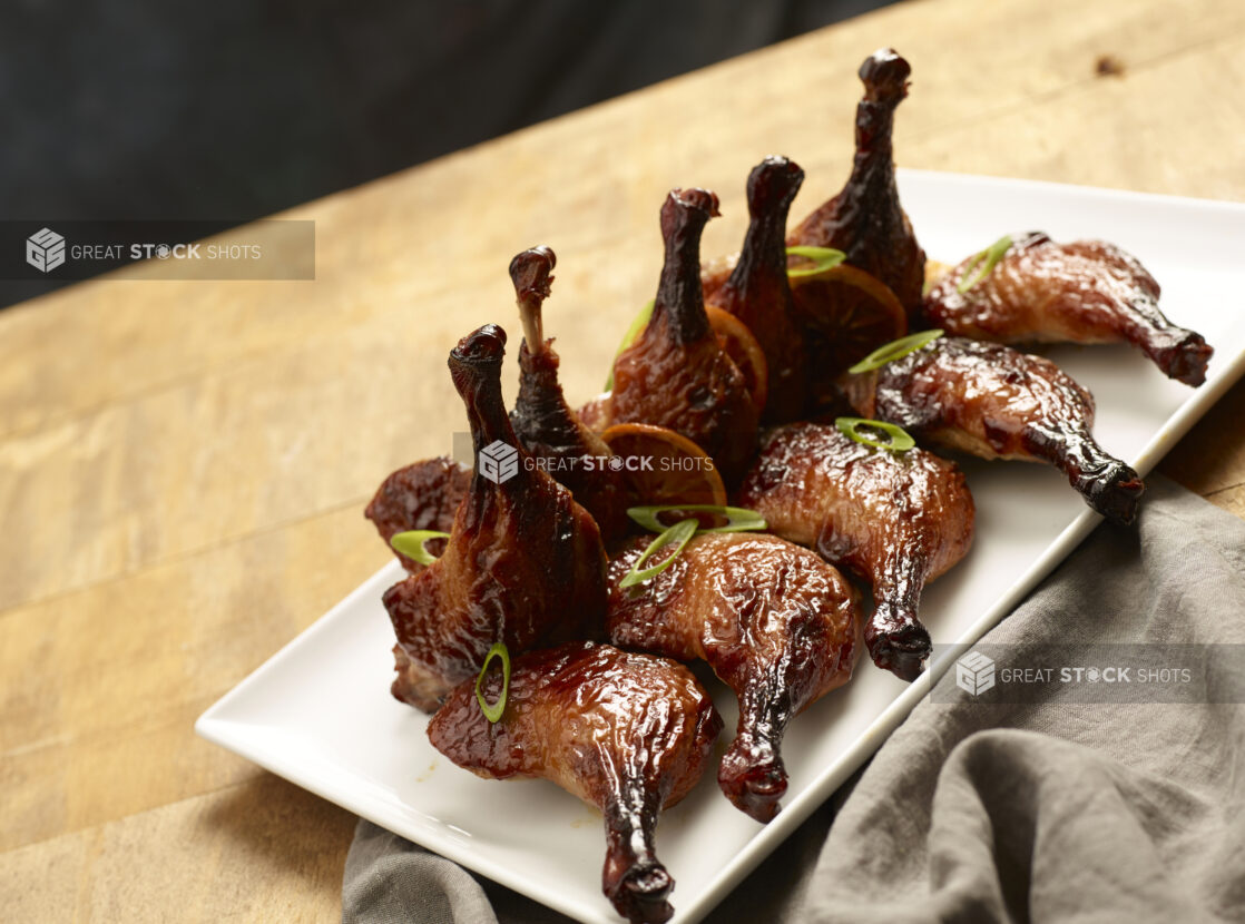 BBQ'd chicken legs, in an elegant display, garnished with green onions and roasted orange slices on a rectangular white ceramic platter