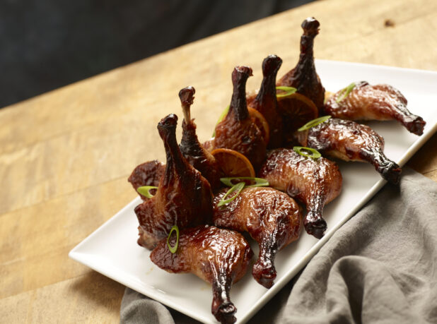 BBQ'd chicken legs, in an elegant display, garnished with green onions and roasted orange slices on a rectangular white ceramic platter