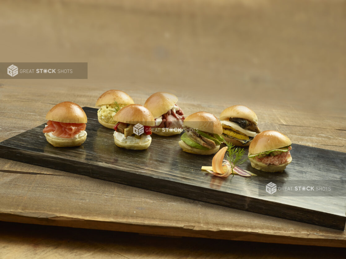 Assorted mini sandwich sliders on on a dark wooden board on a wood table