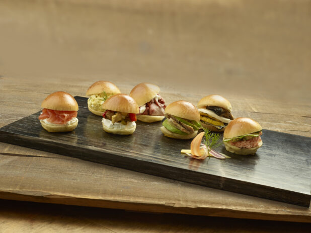 Assorted mini sandwich sliders on on a dark wooden board on a wood table