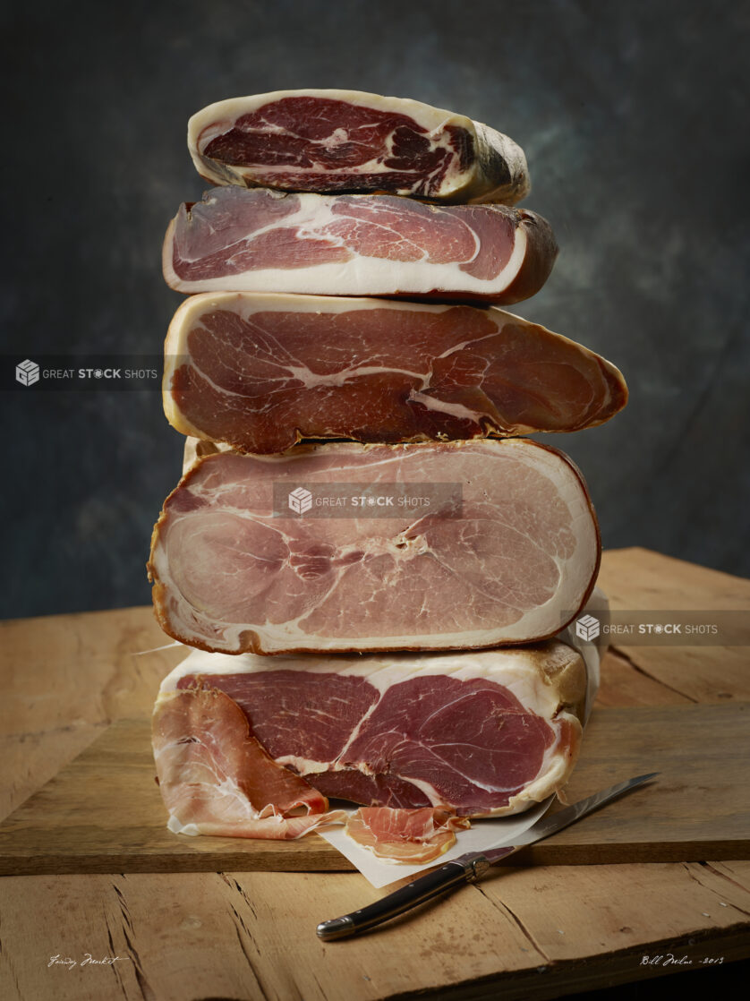 Stacks of cured prosciutto meat with thinly sliced piece at the bottom with a knife on a wooden cutting board