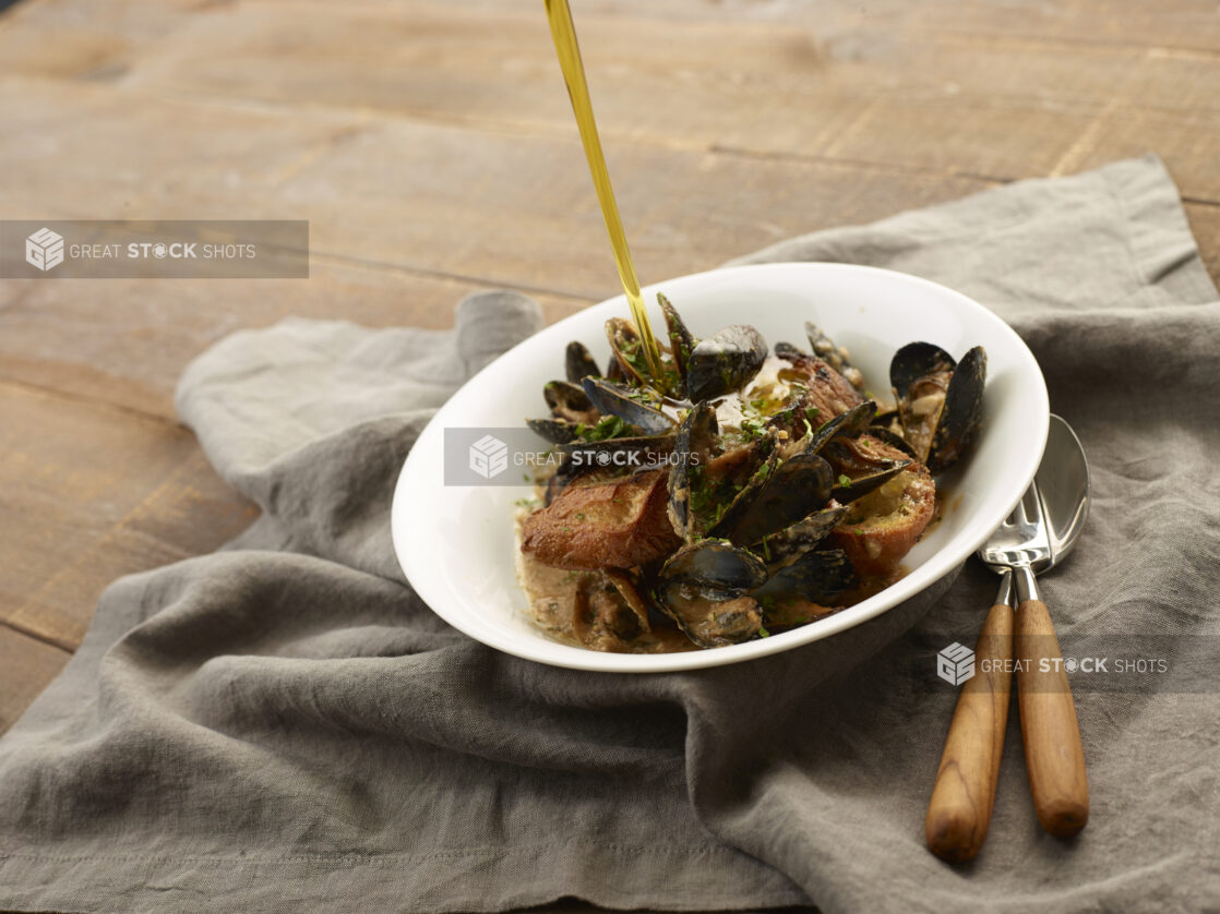 Bowl of mussels on top of toasted italian bread being drizzled with olive oil on a wood table