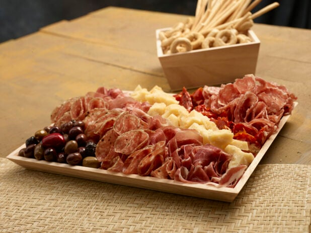 Charcuterie with salami, prosciutto, sundried tomatoes, olives and chunks of parmesan cheese on a wood catering tray with breadsticks and taralli, italian crackers, in a wood catering box on a wooden background