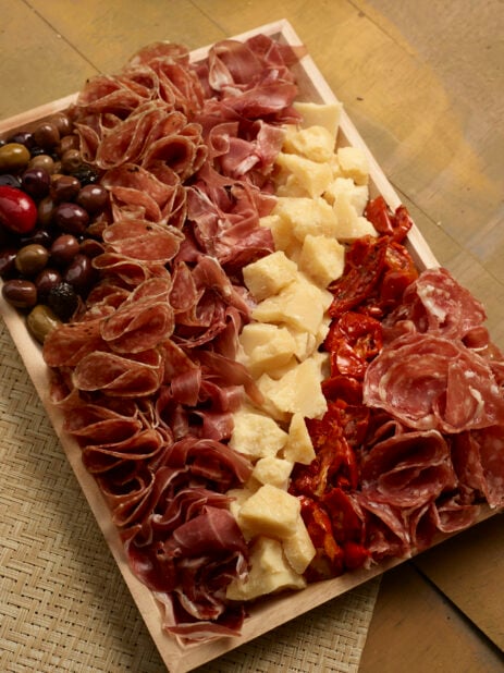 Overhead view of a charcuterie tray with salami, prosciutto, sundried tomatoes, olives and chunks of parmesan cheese on a wood catering tray on a wooden background