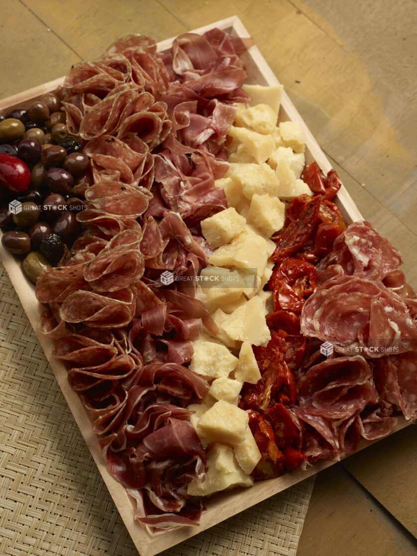 Overhead view of a charcuterie tray with salami, prosciutto, sundried tomatoes, olives and chunks of parmesan cheese on a wood catering tray on a wooden background