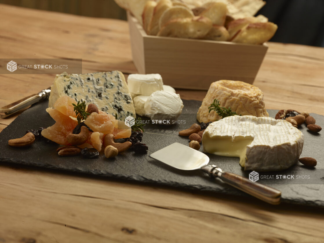 Assorted cheese tray on a slate platter with dried fruit, nuts, cheese knives and a square wood catering box with assorted breads