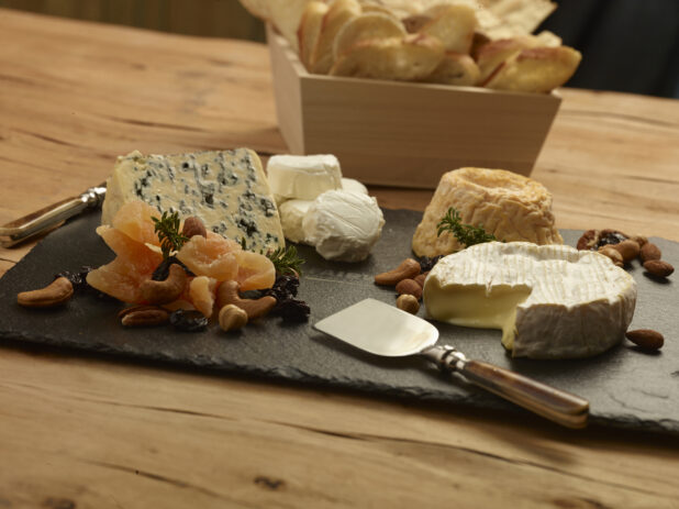 Assorted cheese tray on a slate platter with dried fruit, nuts, cheese knives and a square wood catering box with assorted breads