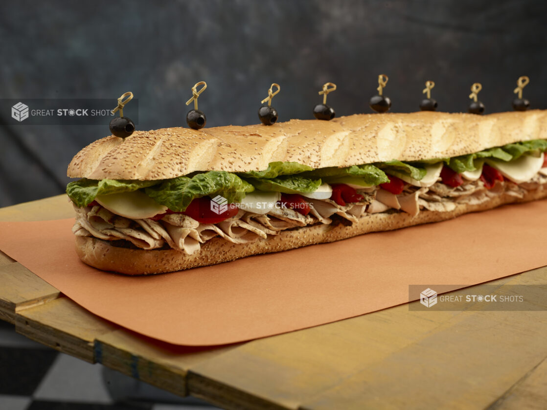 Giant submarine sandwich with sliced roasted chicken, roasted red peppers, cheese and lettuce with bamboo skewers with black olives placed on top of the sandwich