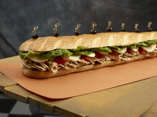 Giant submarine sandwich with sliced roasted chicken, roasted red peppers, cheese and lettuce with bamboo skewers with black olives placed on top of the sandwich