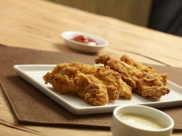 White platter of four chicken fingers with two dipping sauces on a wood table