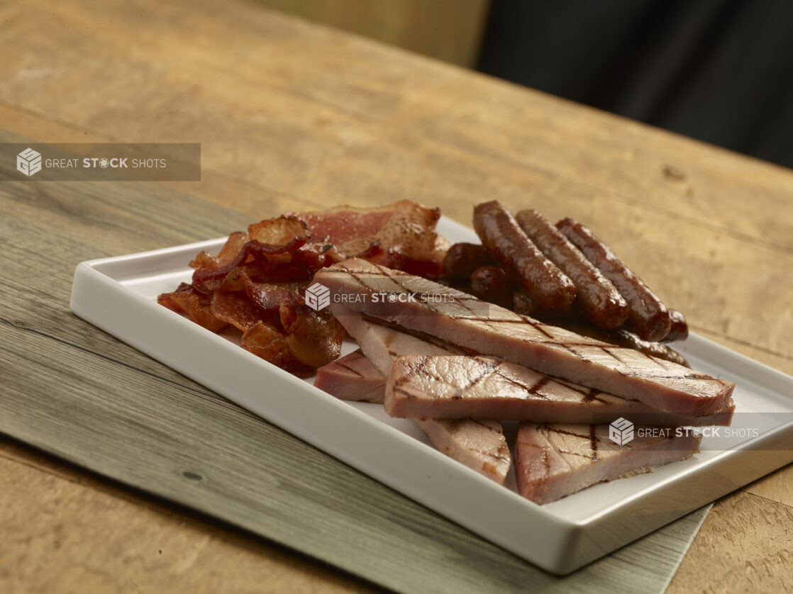 White plate of bacon, grilled ham slices and breakfast sausages on a wood table