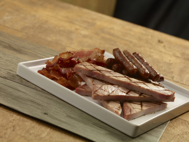 White plate of bacon, grilled ham slices and breakfast sausages on a wood table