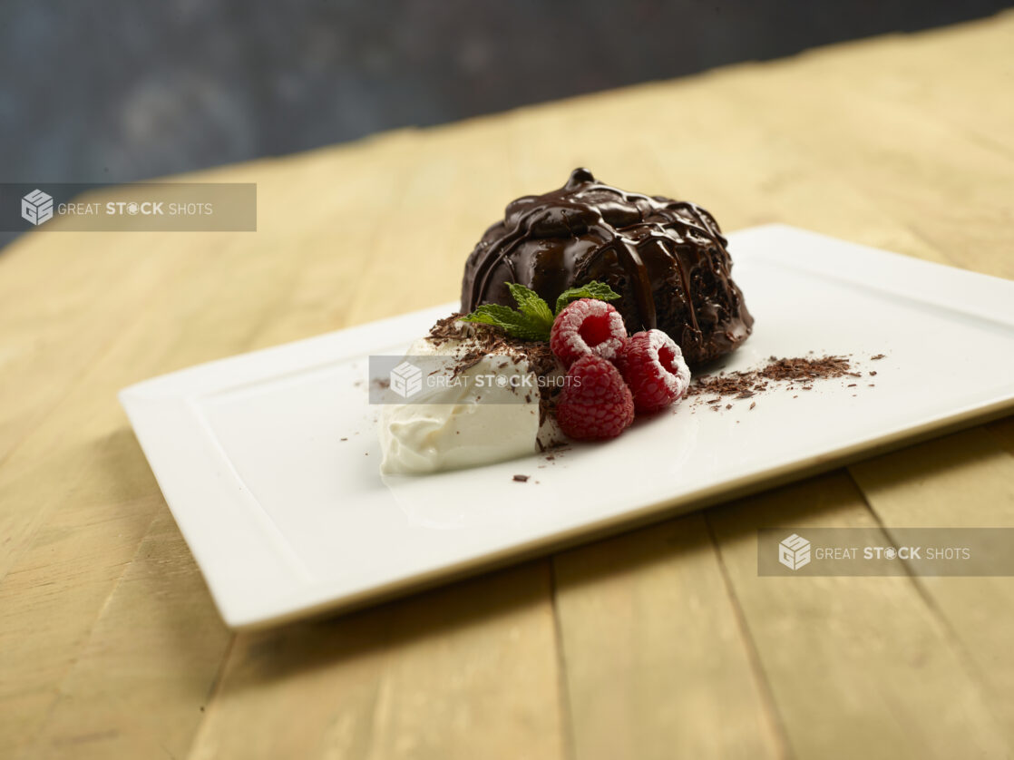 Chocolate lava cake with chocolate drizzle, fresh raspberries, whipped cream and a sprig of mint on a white square plate on a wooden background