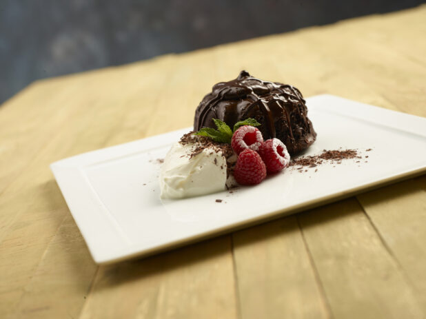 Chocolate lava cake with chocolate drizzle, fresh raspberries, whipped cream and a sprig of mint on a white square plate on a wooden background