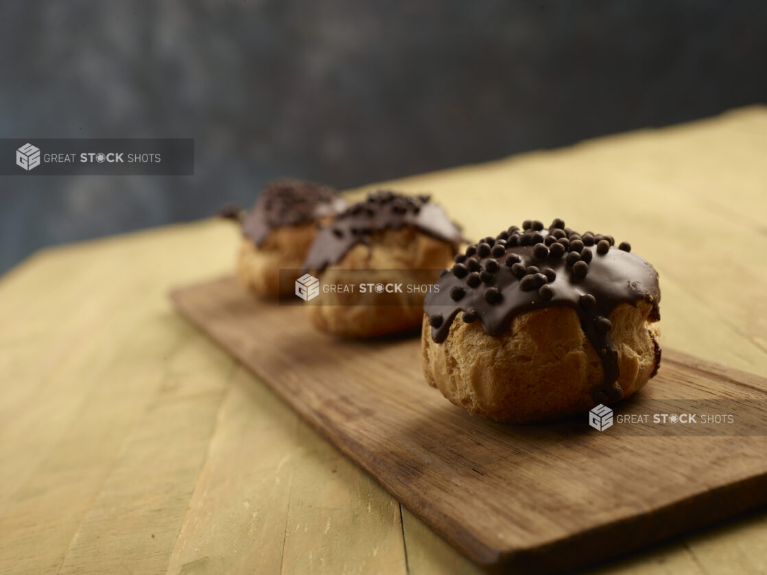 Three chocolate dipped cream puffs with chocolate topping on a wooden board on a wood table