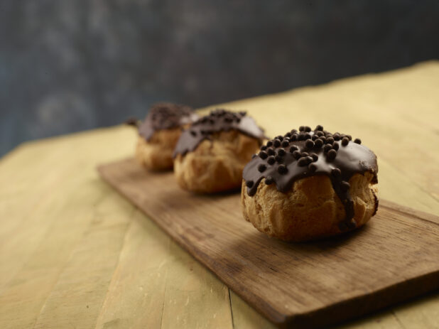 Three chocolate dipped cream puffs with chocolate topping on a wooden board on a wood table