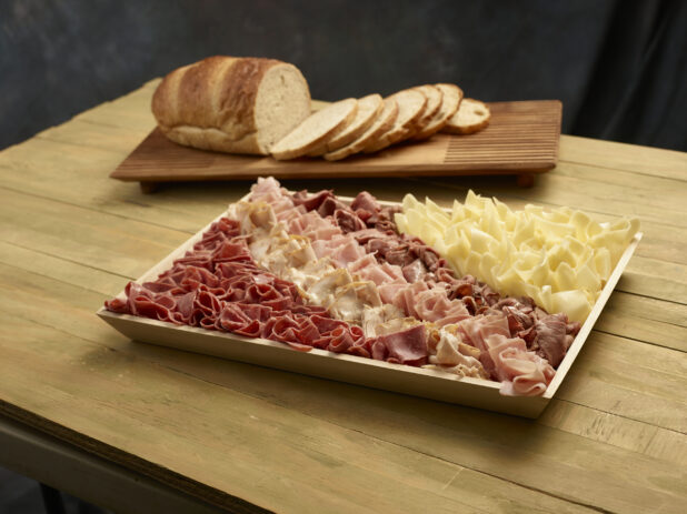 Deli meat and cheese tray on a wood catering tray with light rye bread, half sliced, half unsliced, on wooden board on a wooden background