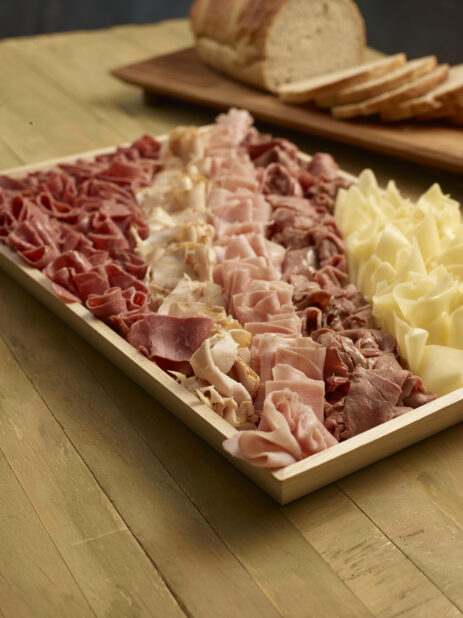 Deli meat and cheese tray on a wood catering tray with light rye bread, half sliced, half unsliced, on wooden board on a wooden background, close-up