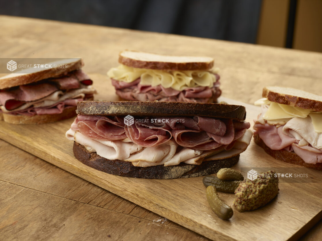 Four sliced deli meat sandwiches on rye bread with gherkin pickles and grainy mustard on the side of a wood board