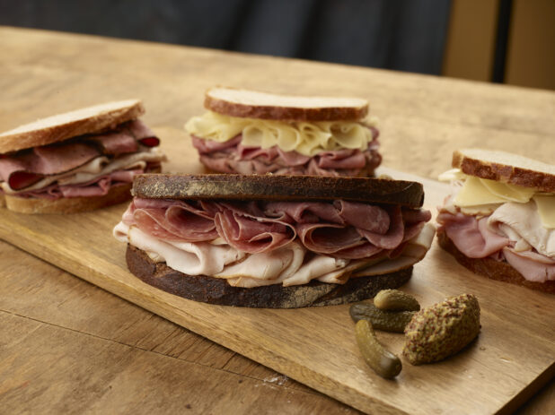 Four sliced deli meat sandwiches on rye bread with gherkin pickles and grainy mustard on the side of a wood board