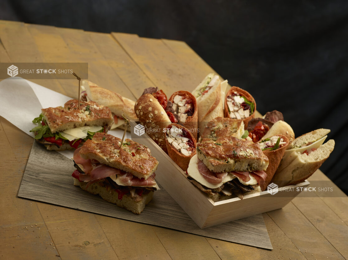 Assorted artisanal sandwiches and wraps in and beside a wood catering box on a wooden background