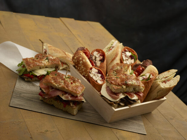 Assorted artisanal sandwiches and wraps in and beside a wood catering box on a wooden background