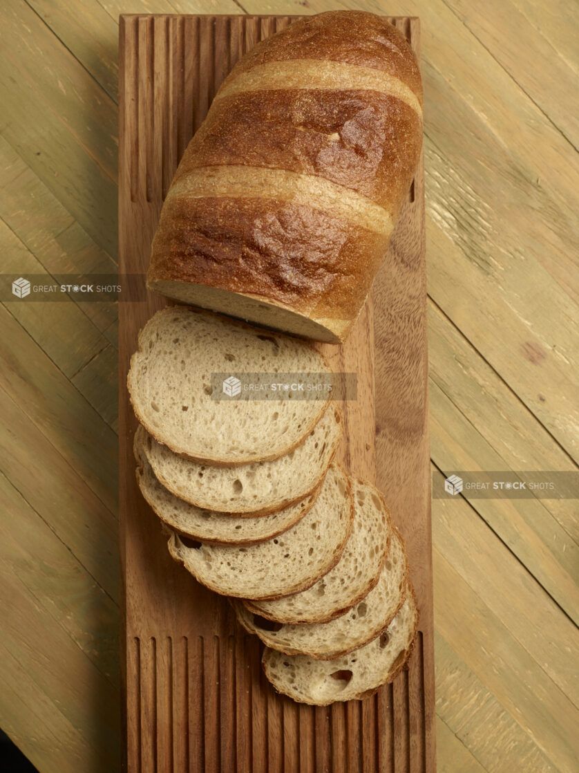 Loaf of light rye bread, half sliced, half unsliced, on wooden board on a wooden background, overhead view