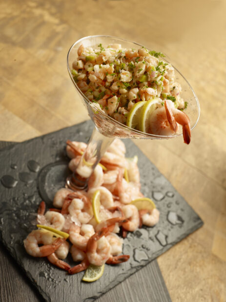 Martini glass filled with shrimp ceviche with whole cooked shrimp and lemon slices around the base on a slate board
