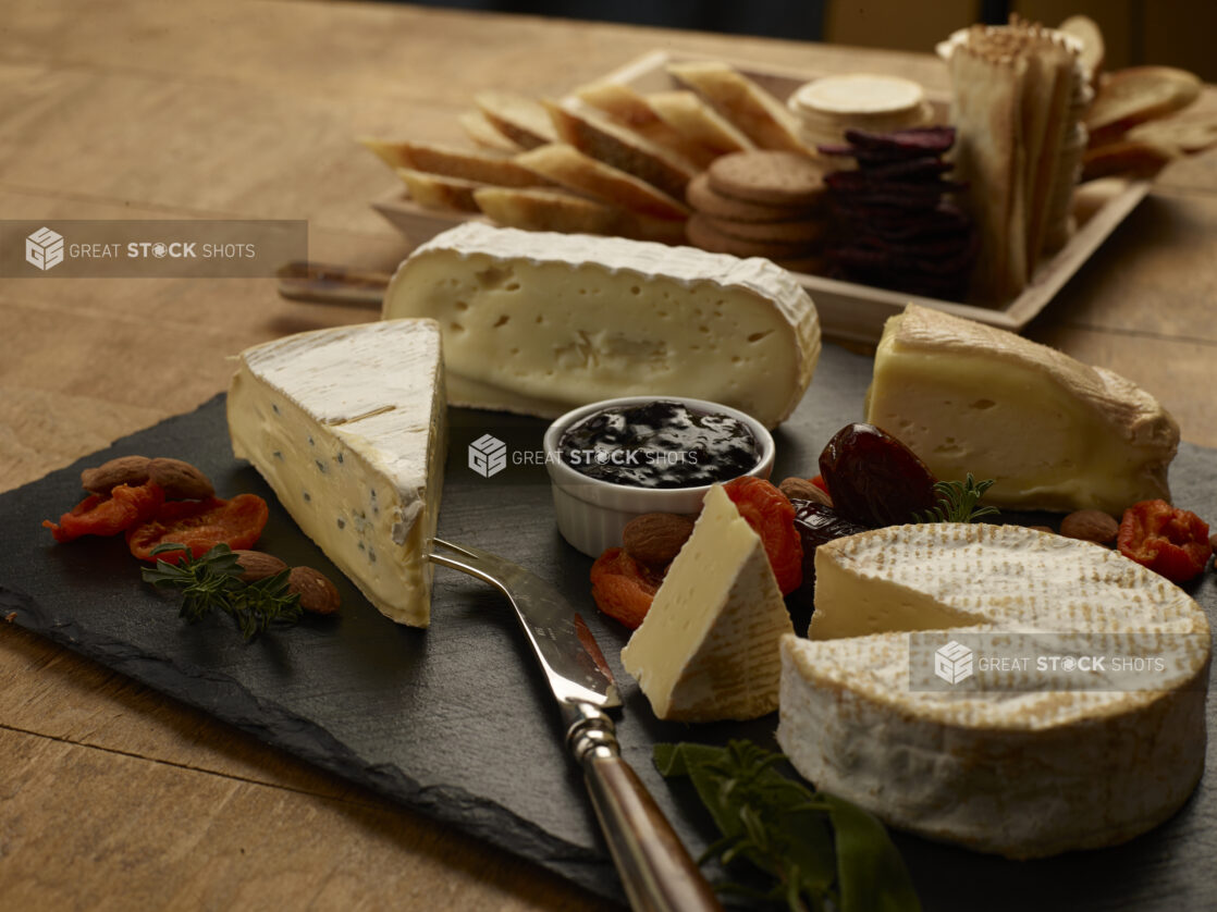 Various cheeses on a slate board with jam, dried fruits and nuts with crackers in the background