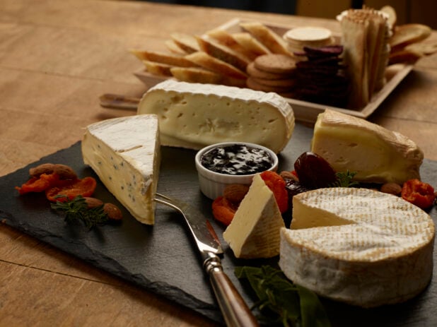Various cheeses on a slate board with jam, dried fruits and nuts with crackers in the background