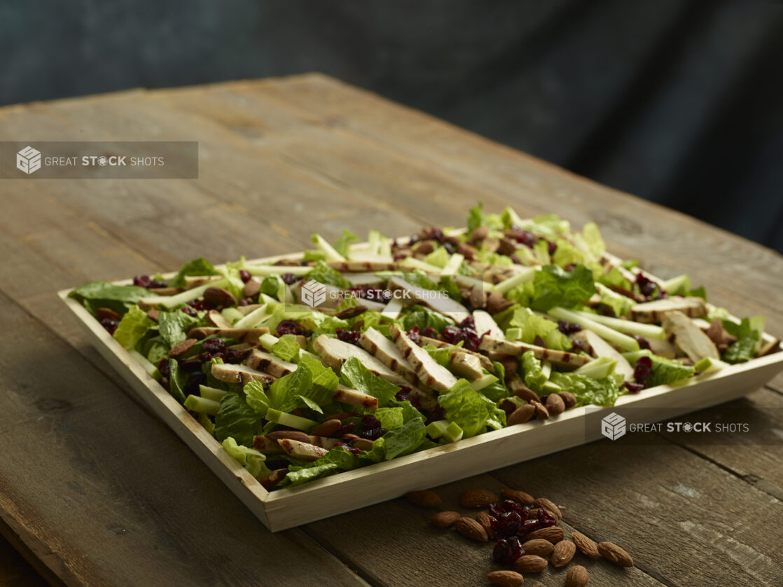 Wood catering tray of grilled chicken salad with green apple, almonds and dried cranberries on a wood table