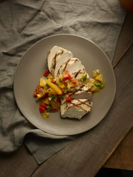 Overhead of sliced grilled chicken breast topped with an orange and pepper sauce on a wood table