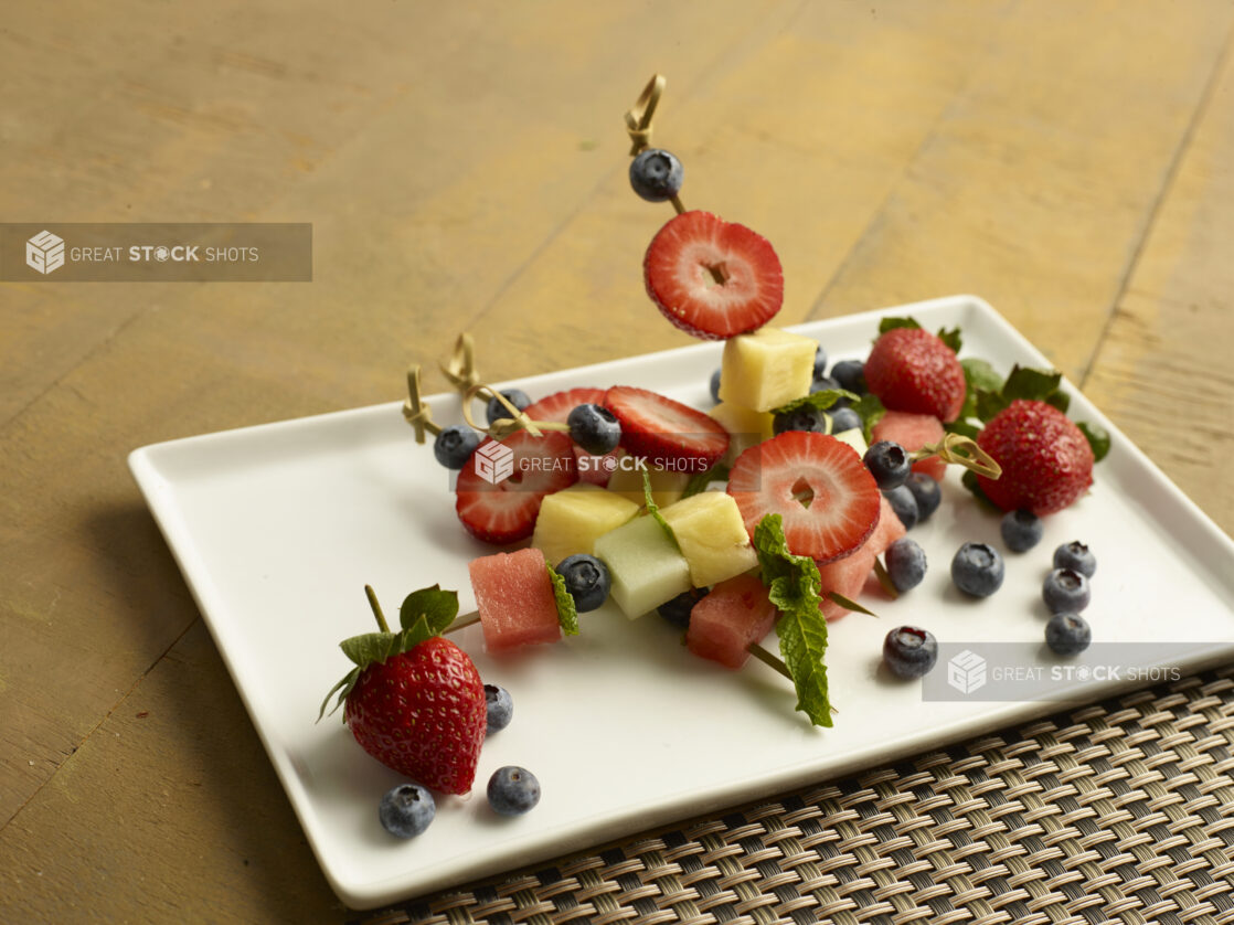 Mixed fruit skewered with a bamboo toothpick on a white plate on a wood table