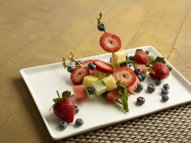 Mixed fruit skewered with a bamboo toothpick on a white plate on a wood table