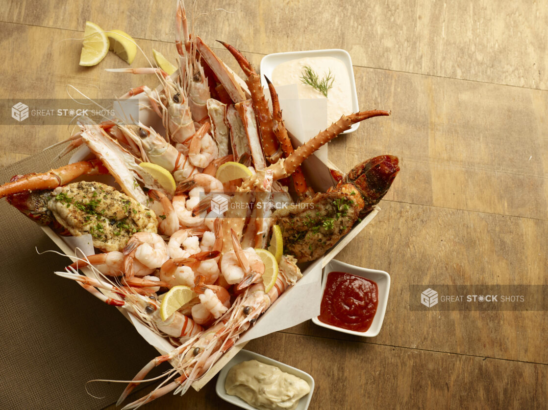 Seafood platter of crab legs, butterflied lobster tail, tiger prawns and shrimp in a wood tray with lemon wedges with dips on a wood table