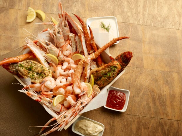 Seafood platter of crab legs, butterflied lobster tail, tiger prawns and shrimp in a wood tray with lemon wedges with dips on a wood table