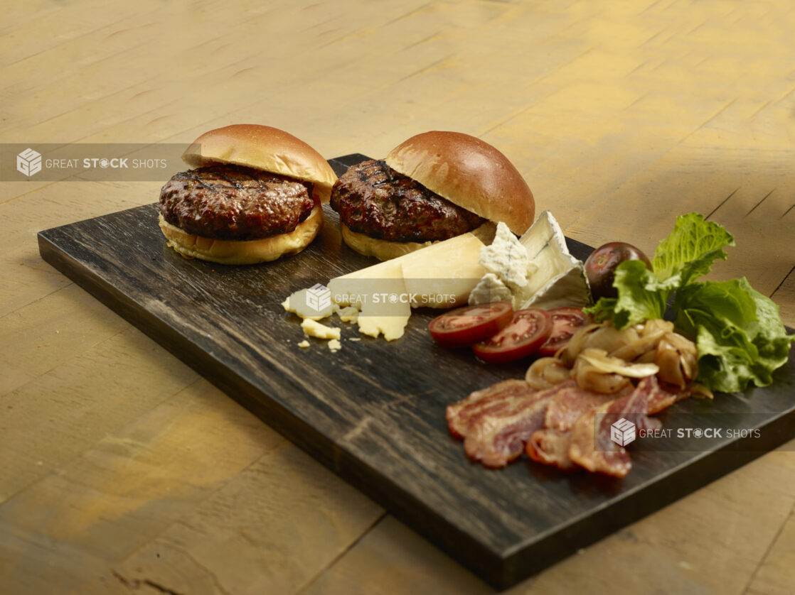 Two naked hamburgers sitting on a wooden board with toppings of parmesan and blue cheese, sliced tomatoes, caramelized onion, bacon, lettuce and tomato