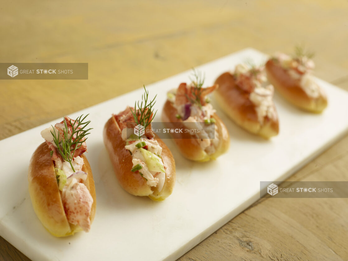 Five mini lobster rolls with fresh dill on a white marble slab on a wood table