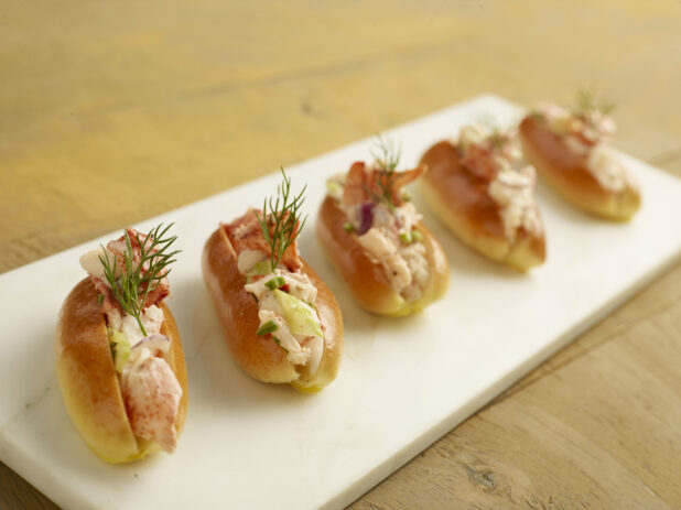 Five mini lobster rolls with fresh dill on a white marble slab on a wood table