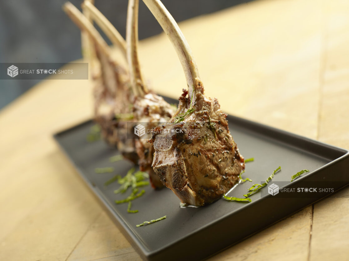 Four grilled lamb chops standing on a black platter topped with fresh basil on a wood table