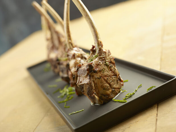Four grilled lamb chops standing on a black platter topped with fresh basil on a wood table