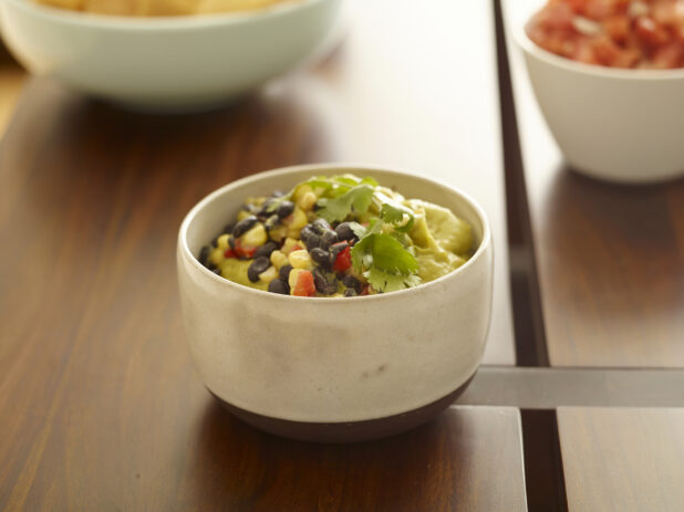 Small bowl of guacamole topped with corn, black beans, diced red pepper and cilantro on a wood table