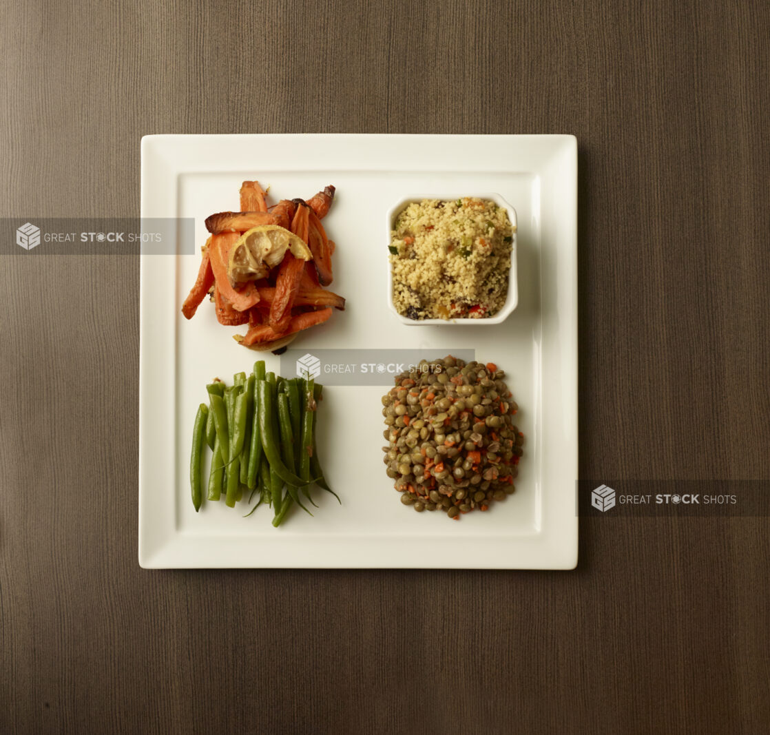 Overhead of four side dishes of roasted carrots, couscous salad, green beans and french lentils on a white plate on a wood table