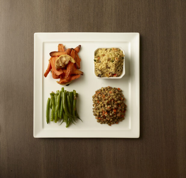 Overhead of four side dishes of roasted carrots, couscous salad, green beans and french lentils on a white plate on a wood table