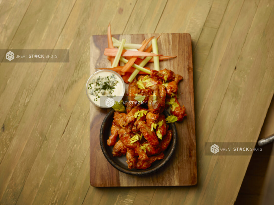 Overhead of sauced chicken wings on a wood board with a side carrots, celery and blue cheese dip on a wood table