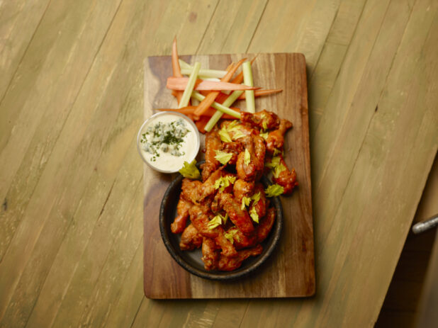 Overhead of sauced chicken wings on a wood board with a side carrots, celery and blue cheese dip on a wood table