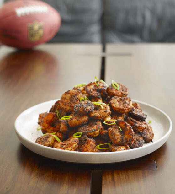 Plate of blackened barbecued shrimp topped with green onion in a home on a wood table with a football