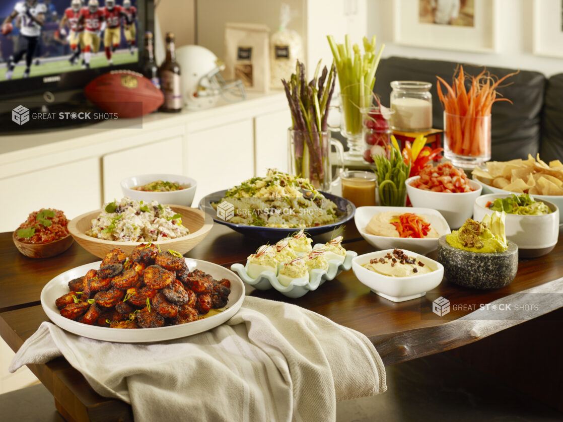 Assortment of dips and appetizers on a wood table in a home watching football on the television