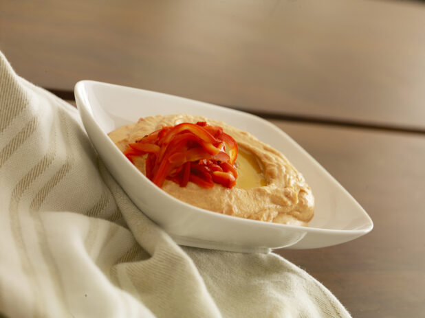 White bowl of roasted red pepper hummas topped with roasted red peppers and olive oil on a wood table