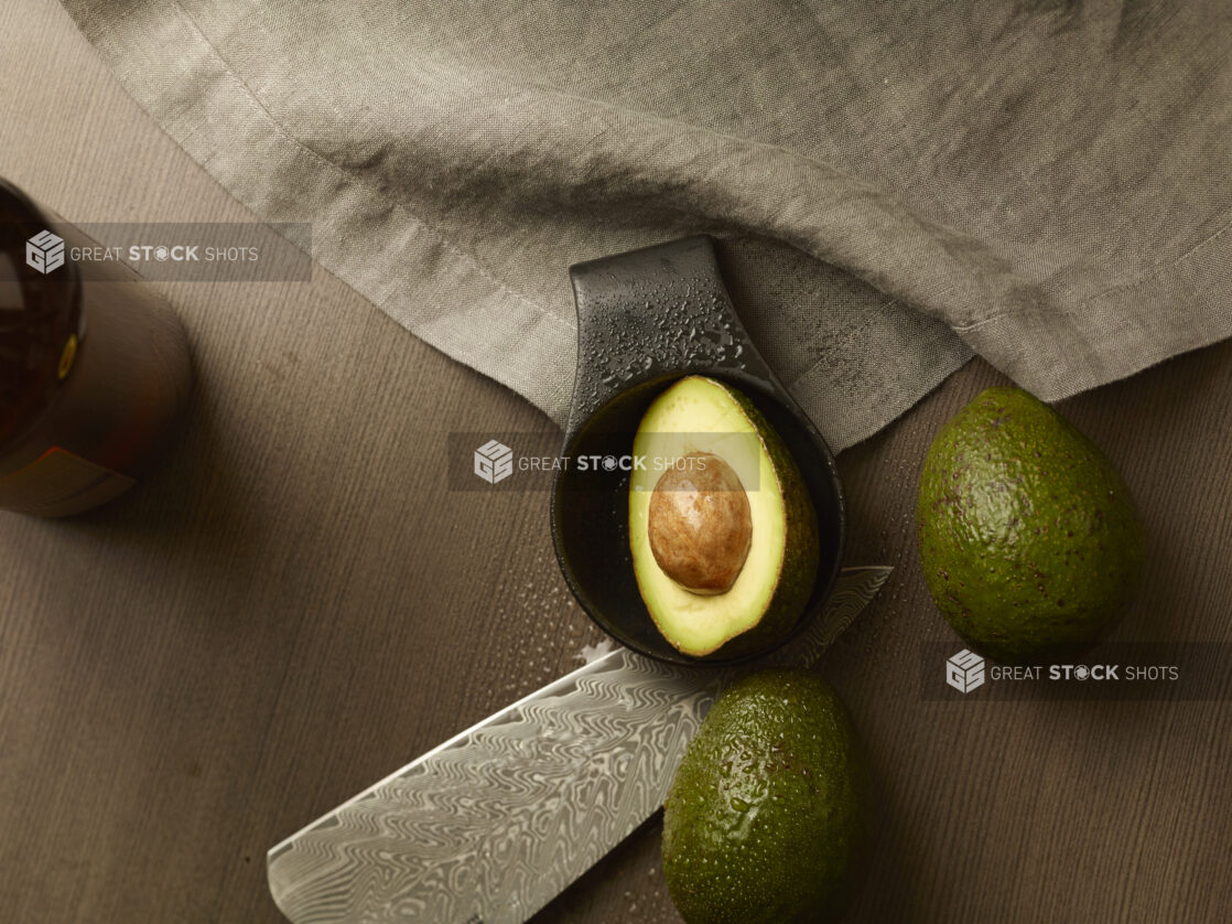 Overhead of an avocado cut in half in a dish with two other avocados and a knife around it on a wood table